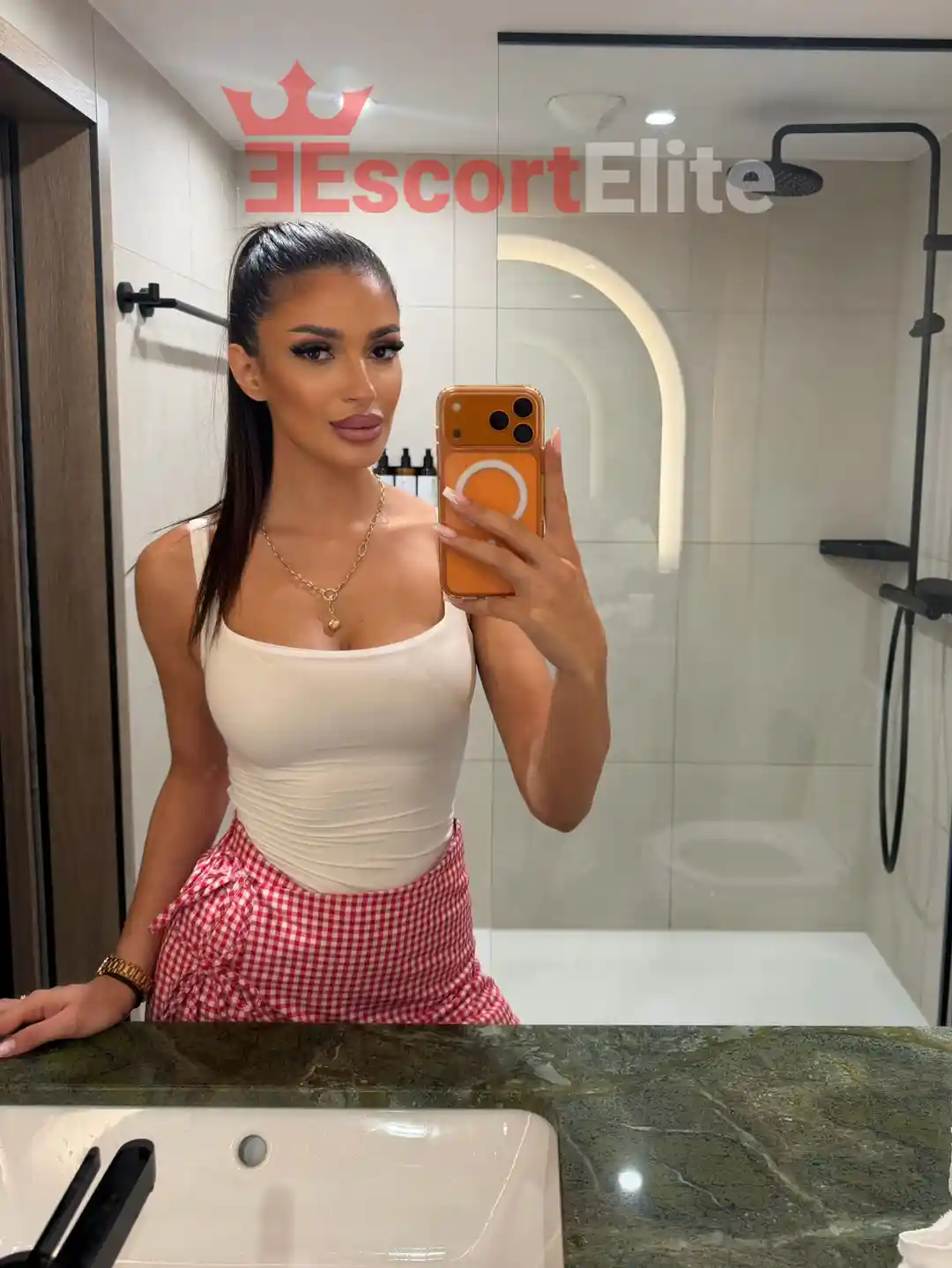 Escort Wien Kassandra im weißen Top mit rotem Karo-Rock, Spiegelselfie im Badezimmer